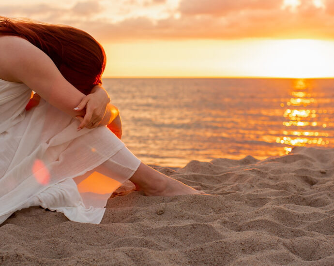 Profile of a woman in a long white dress, sitting on the beach at sunset, feet in the sand, with her head down, grieving a loss.