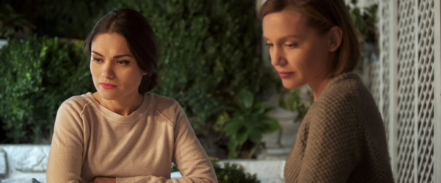 Two ladies, with coffee cups in front of them, sit beside each other at a wooden table on an outdoor patio. Each is looking away, seemingly to avoid a difficult situation or a troubling conversation. Their body language suggests that they are experiencing a communication problem after having a personal conflict.