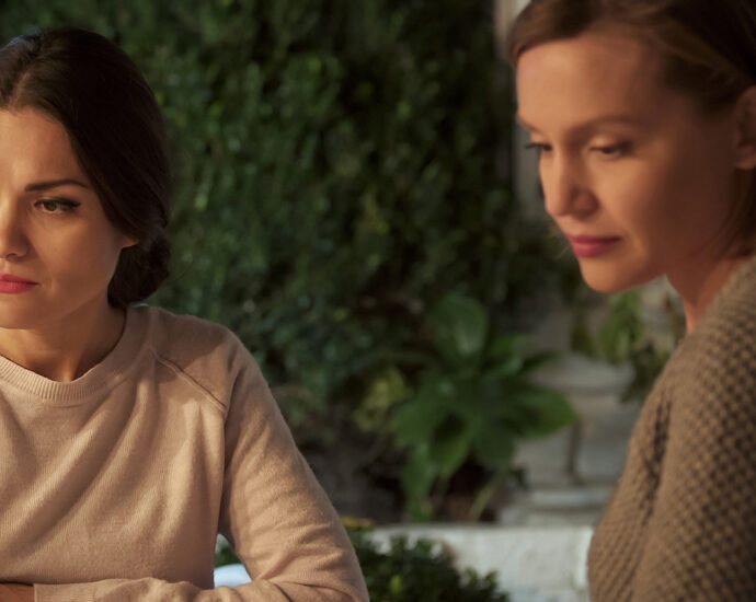 Two ladies, with coffee cups in front of them, sit beside each other at a wooden table on an outdoor patio. Each is looking away, seemingly to avoid a difficult situation or a troubling conversation. Their body language suggests that they are experiencing a communication problem after having a personal conflict.