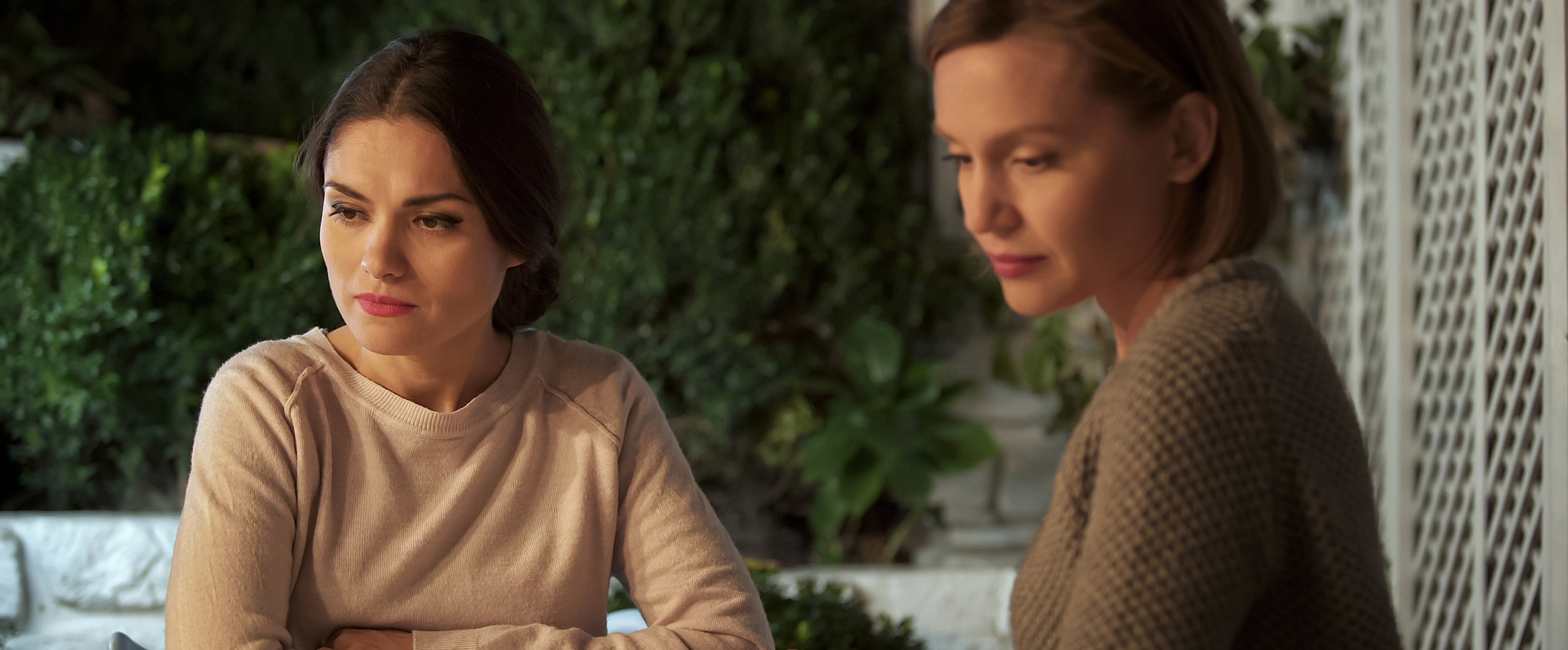 Two ladies, with coffee cups in front of them, sit beside each other at a wooden table on an outdoor patio. Each is looking away, seemingly to avoid a difficult situation or a troubling conversation. Their body language suggests that they are experiencing a communication problem after having a personal conflict.