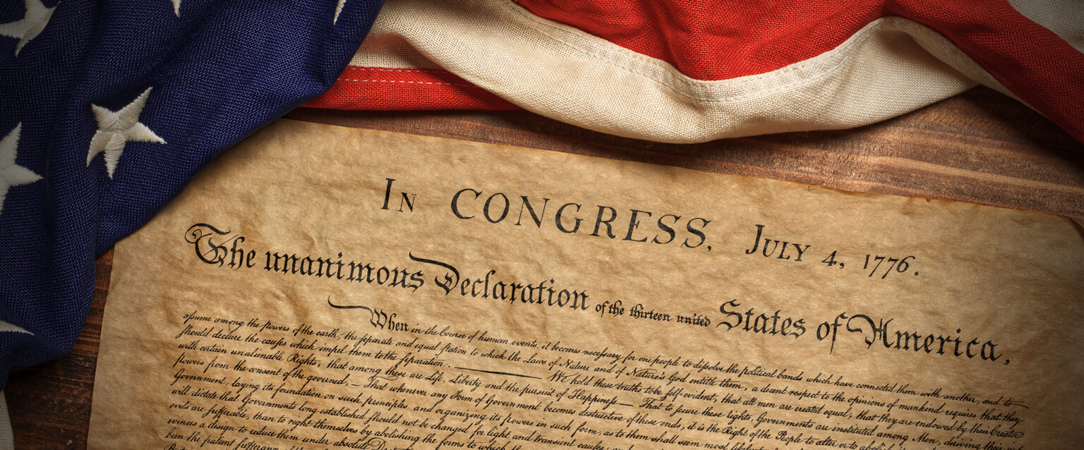 The United States Declaration of Independence, lying on a wooden table, with an American flag casually draped across the table above it.