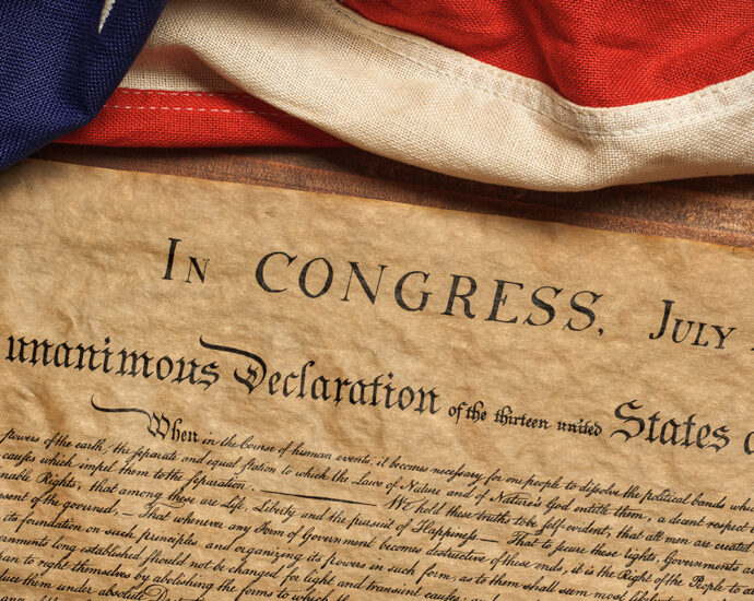 The United States Declaration of Independence, lying on a wooden table, with an American flag casually draped across the table above it.