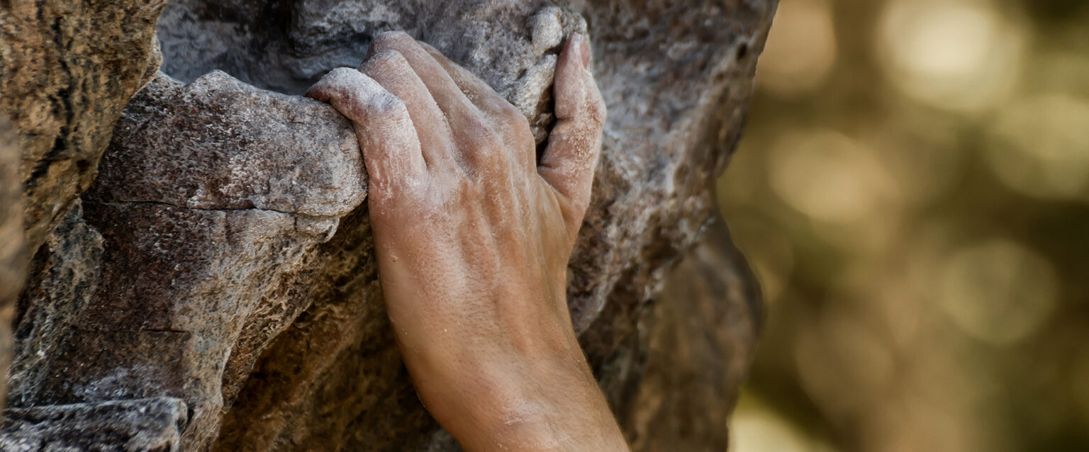 A close-up view of a rock climber's hand gripping a handhold on a natural cliff outcropping