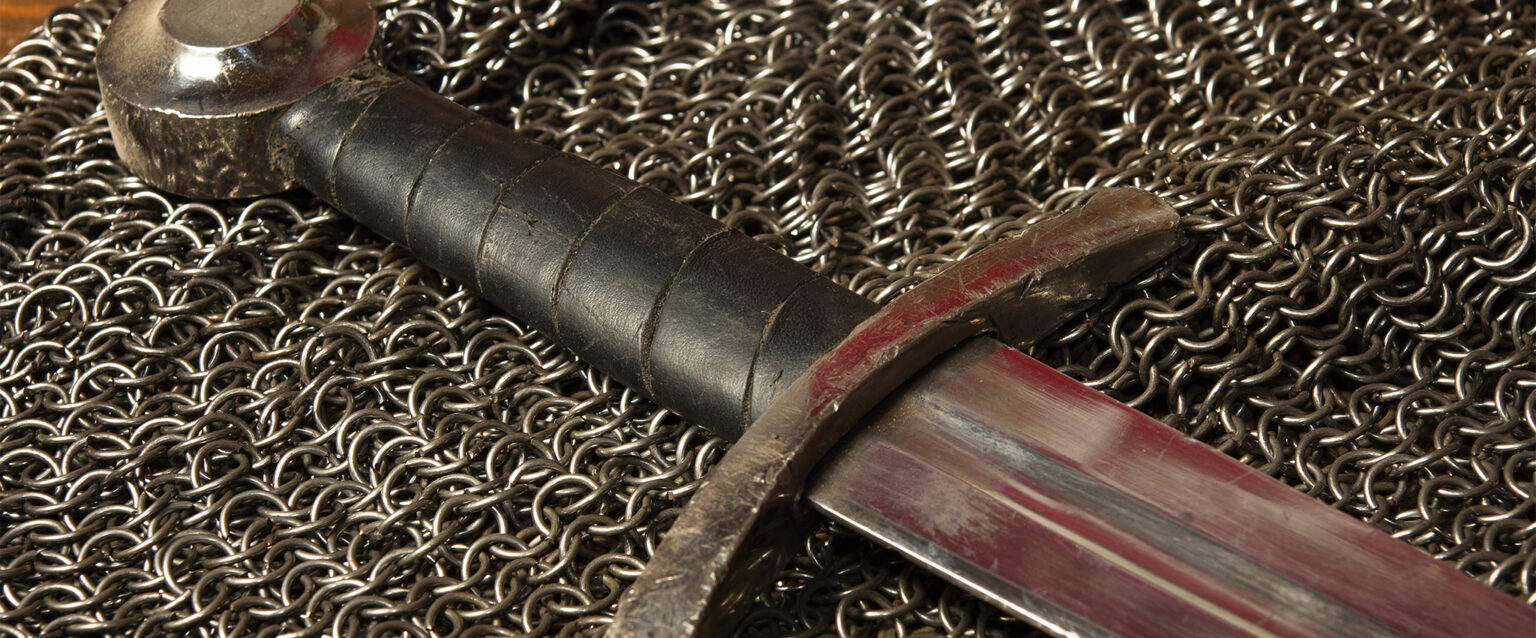 A still life photo of a medieval sword and hauberk. A steel sword, with leather-wrapped handle lies atop a piece of chainmail armor. A red reflection in the blade and hilt of the well-worn sword appears as though it was bloodied in battle before being laid down.
