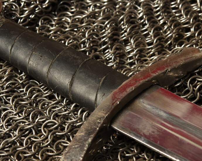 A still life photo of a medieval sword and hauberk. A steel sword, with leather-wrapped handle lies atop a piece of chainmail armor. A red reflection in the blade and hilt of the well-worn sword appears as though it was bloodied in battle before being laid down.