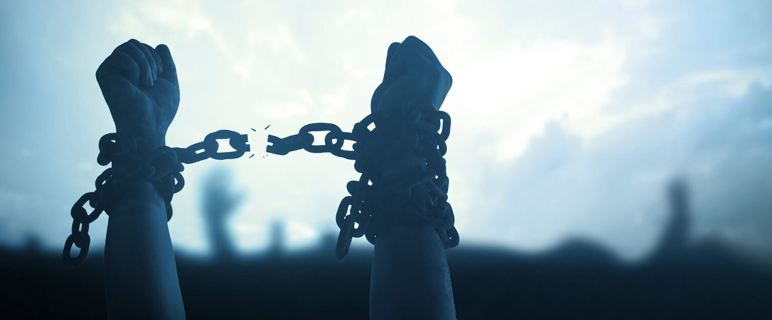 A cool-toned photo of human arms, with chains wrapped around their wrists, hands raised above a crowd. The chain between their wrists is breaking, expressing the message of gained freedom through fight.