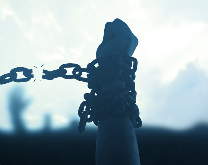 A cool-toned photo of human arms, with chains wrapped around their wrists, hands raised above a crowd. The chain between their wrists is breaking, expressing the message of gained freedom through fight.
