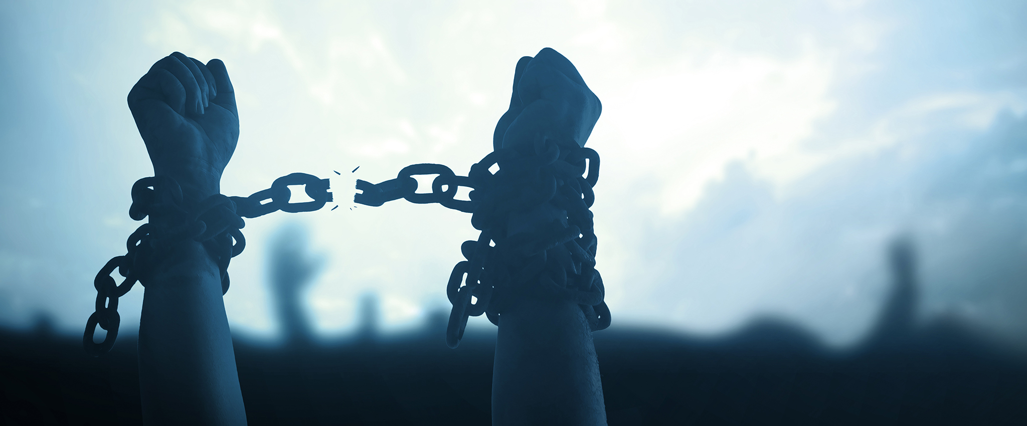 A cool-toned photo of human arms, with chains wrapped around their wrists, hands raised above a crowd. The chain between their wrists is breaking, expressing the message of gained freedom through fight.
