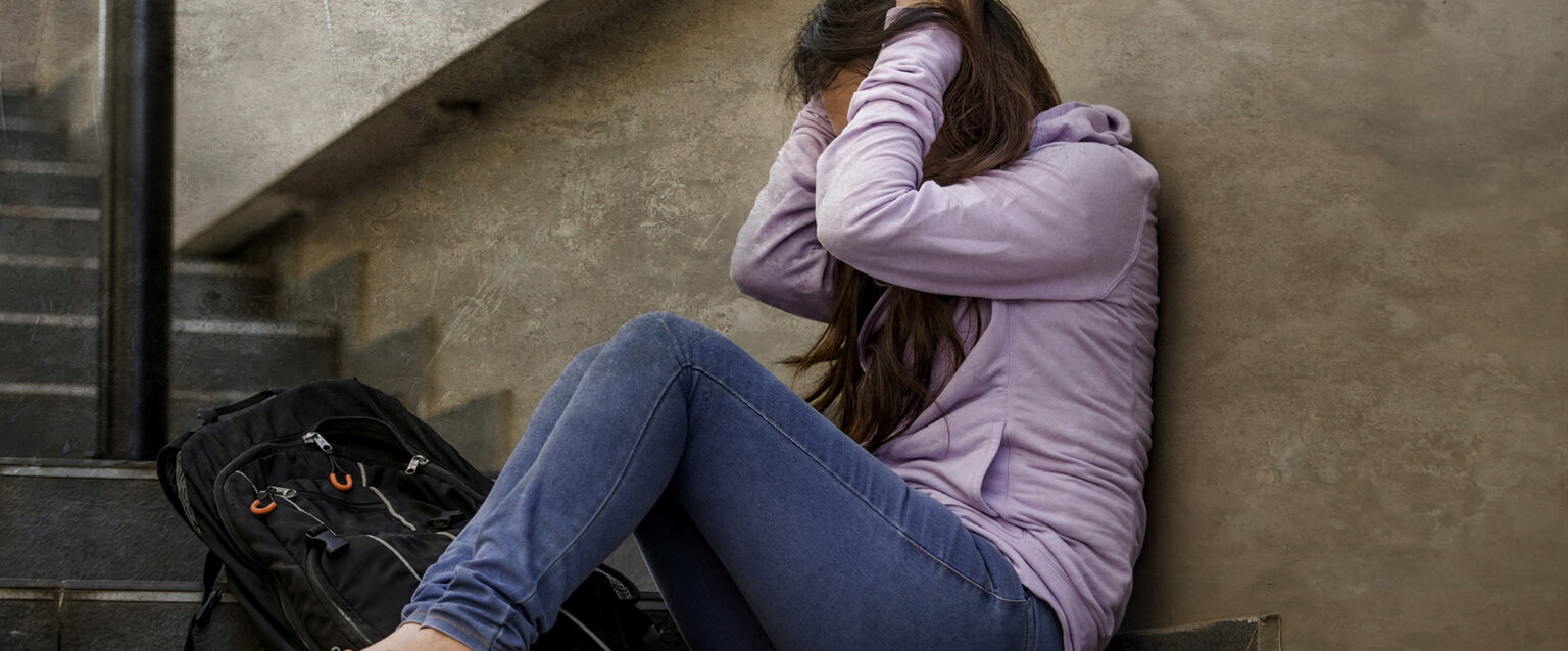 Side view of an upset female student wearing jeans and a lavender hoodie. Sitting on an outdoor cement stairway, she leans against the wall beside it, her hands holding the side of her head in anguish.
