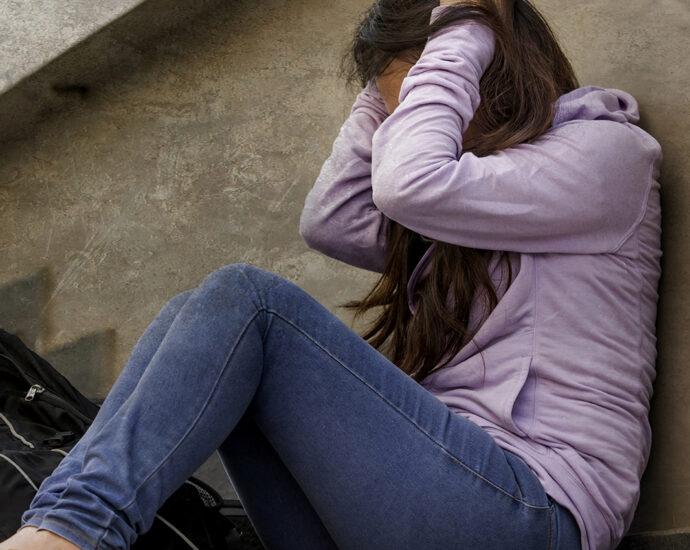 Side view of an upset female student wearing jeans and a lavender hoodie. Sitting on an outdoor cement stairway, she leans against the wall beside it, her hands holding the side of her head in anguish.