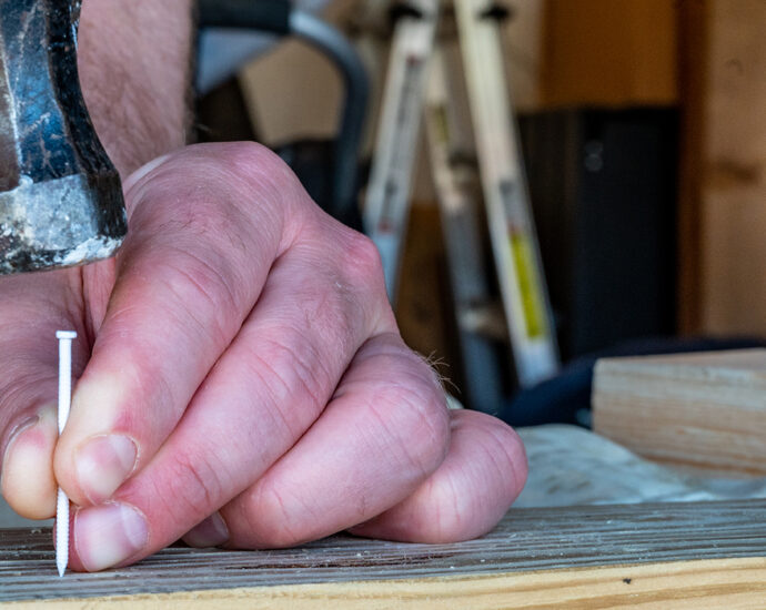 A man preparing to drive a white, painted finishing nail into a board with a hammer while his left hand supports the nail. A ladder, shop benches and tools are visible in the background.