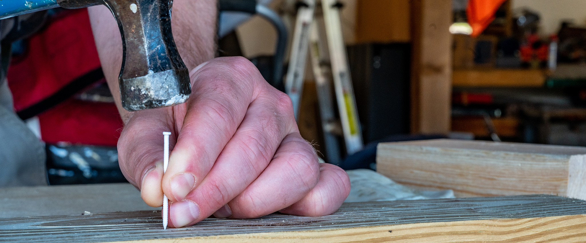 A man preparing to drive a white, painted finishing nail into a board with a hammer while his left hand supports the nail. A ladder, shop benches and tools are visible in the background.