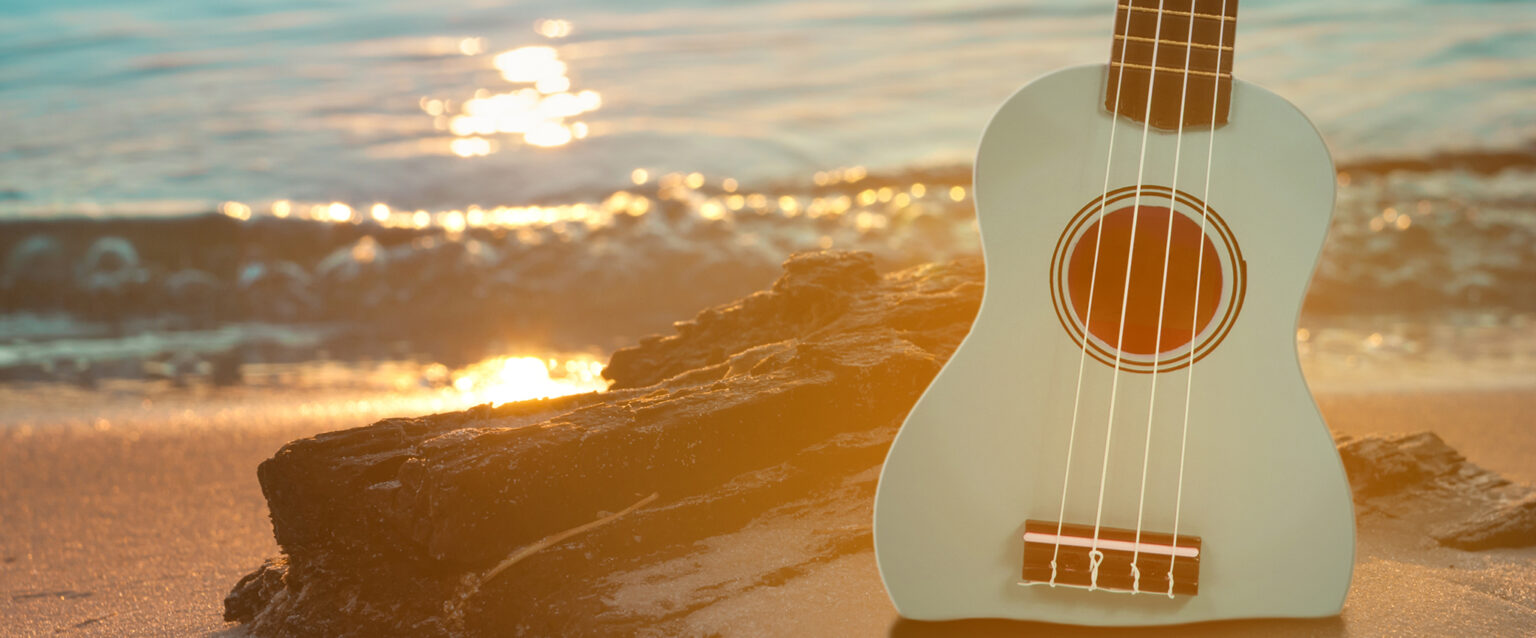A powder blue ukulele sits upright in the sand on a rocky beach with clear blue water and small waves.