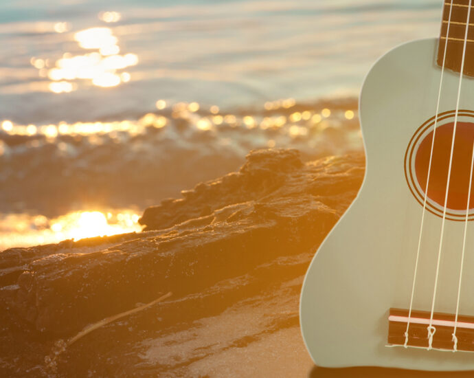 A powder blue ukulele sits upright in the sand on a rocky beach with clear blue water and small waves.