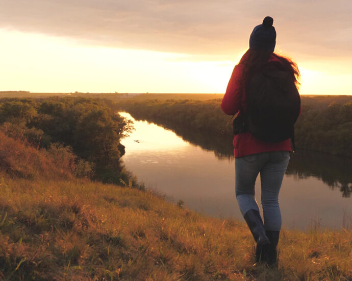 A young girl, wearing jeans, a red shirt, tall boots, a winter hat with a pom-pom, and a black backpack, walks along a grass-covered ridge near a river at sunset.