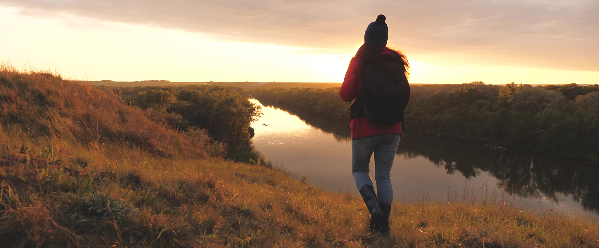 A young girl, wearing jeans, a red shirt, tall boots, a winter hat with a pom-pom, and a black backpack, walks along a grass-covered ridge near a river at sunset.