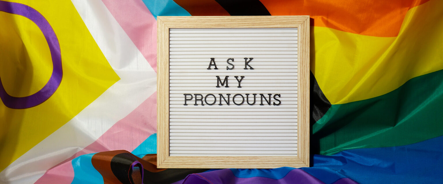 Black, plastic letters spelling out "ASK MY PRONOUNS" on a reusable, wood-framed square sign, nestled on a wrinkled rainbow PRIDE flag.