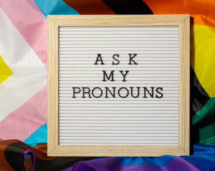 Black, plastic letters spelling out "ASK MY PRONOUNS" on a reusable, wood-framed square sign, nestled on a wrinkled rainbow PRIDE flag.