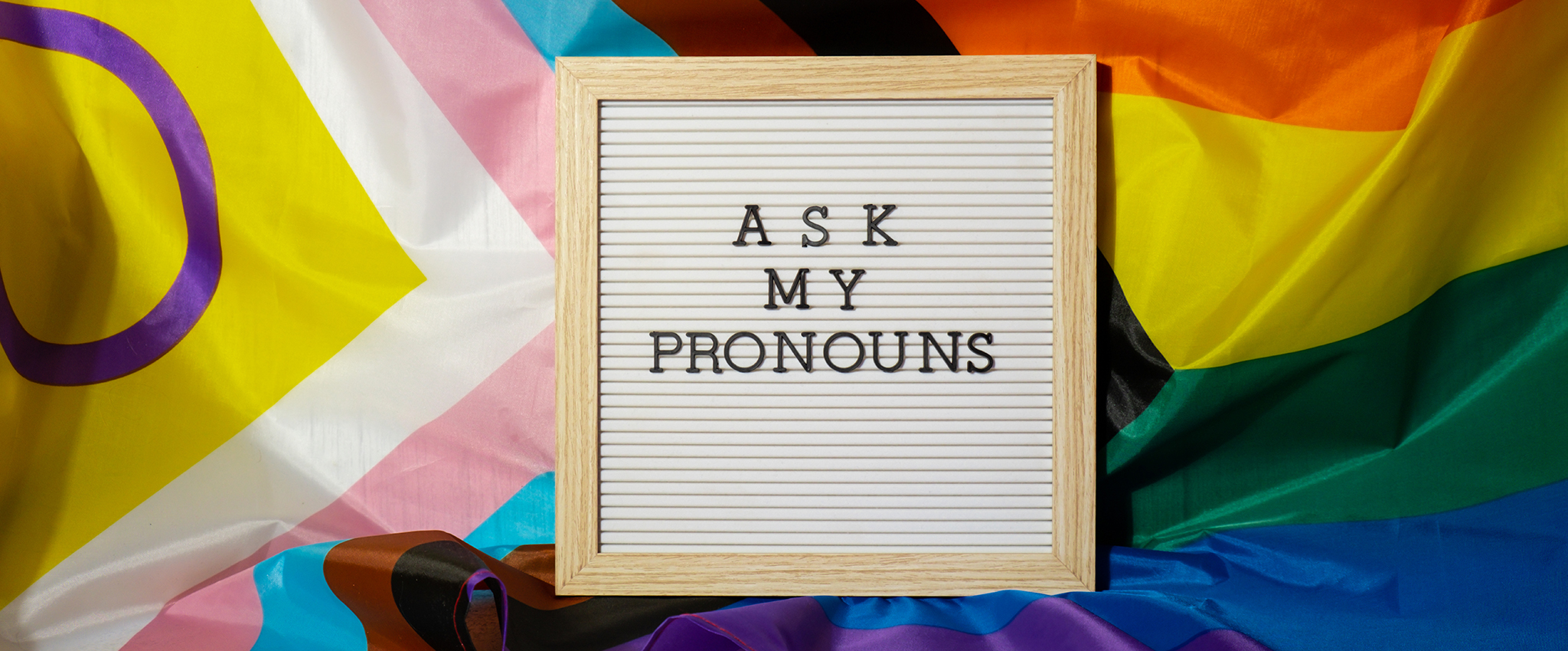 Black, plastic letters spelling out "ASK MY PRONOUNS" on a reusable, wood-framed square sign, nestled on a wrinkled rainbow PRIDE flag.