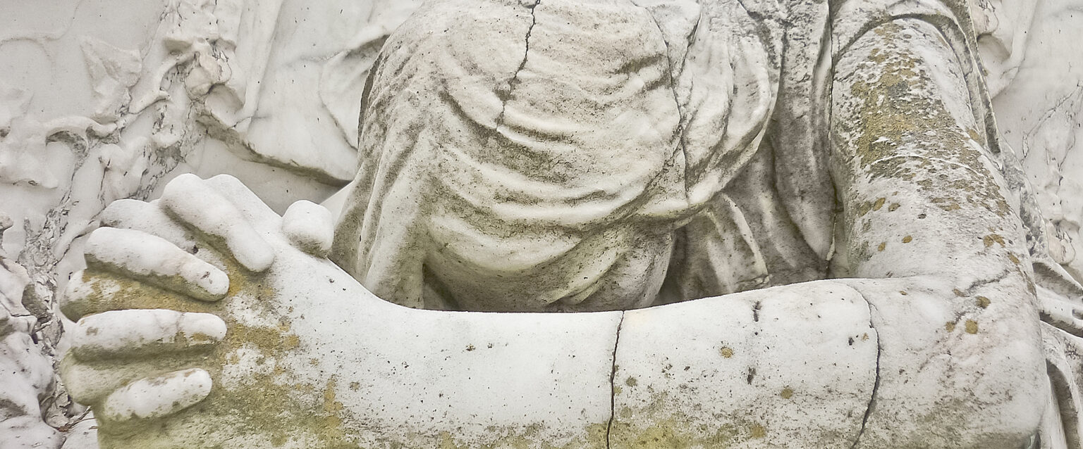 Close-up of a weathered and cracked, white statue of an angel leaning on a table, head down, hands clasped, fingers interlocked, praying. Located in an Uruguayan cemetery.