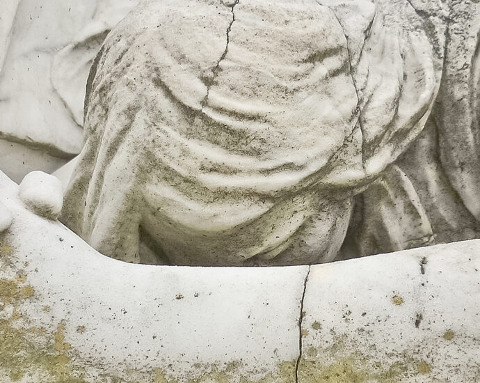 Close-up of a weathered and cracked, white statue of an angel leaning on a table, head down, hands clasped, fingers interlocked, praying. Located in an Uruguayan cemetery.