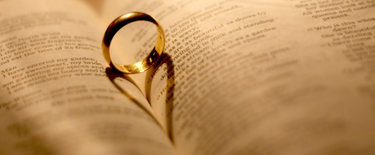 A plain gold wedding Ring sits upright between two curved pages in an open Bible. Back lighting creates a shadow in the shape of a heart, which extends into the crease.