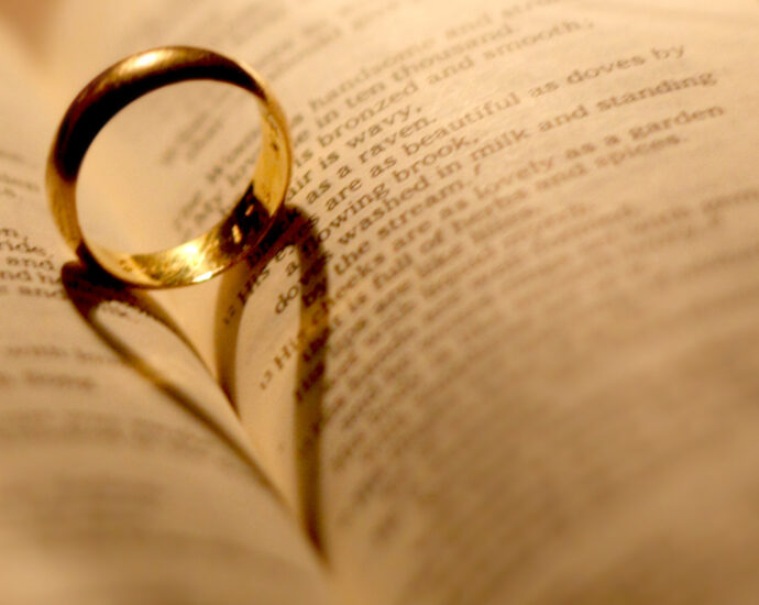 A plain gold wedding Ring sits upright between two curved pages in an open Bible. Back lighting creates a shadow in the shape of a heart, which extends into the crease.