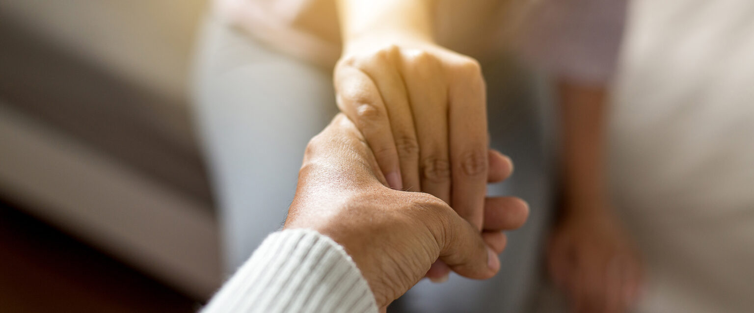 A man, caringly holding his hand out to help a woman, who is sitting on a hospital bed with her hand in his. The main focus is on their hands.