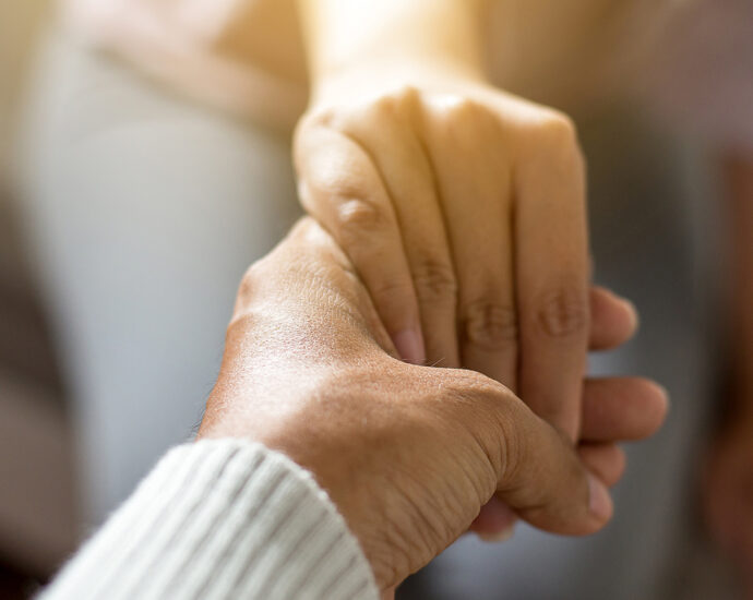 A man, caringly holding his hand out to help a woman, who is sitting on a hospital bed with her hand in his. The main focus is on their hands.