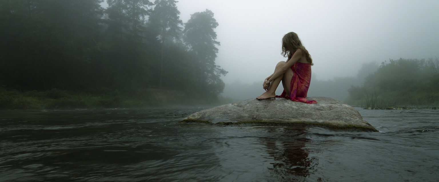 A reclusive girl, in a red dress, sitting alone on a lone rock in the middle of a river, in the early morning