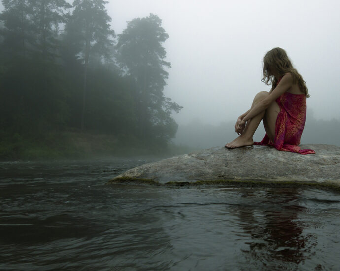 A reclusive girl, in a red dress, sitting alone on a lone rock in the middle of a river, in the early morning