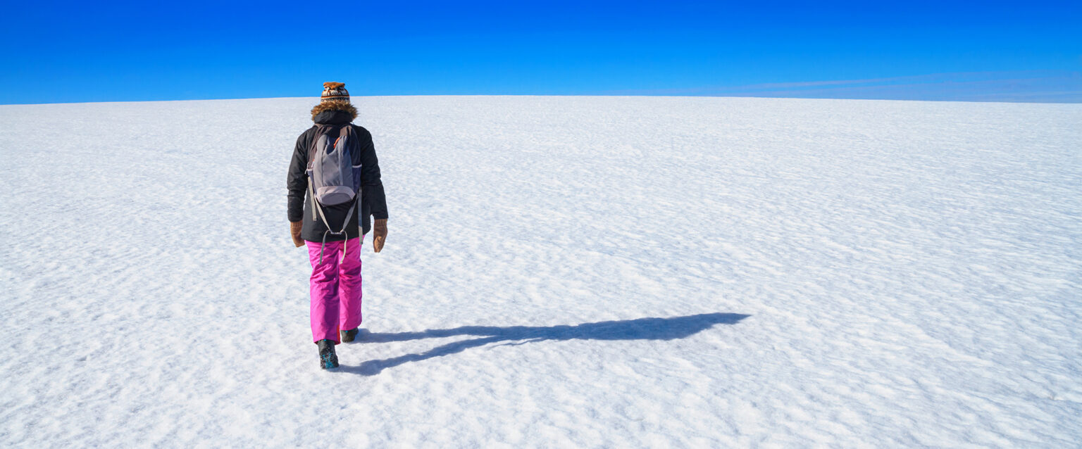An empty, snow-covered, arctic winter landscape; a snowy desert field with a person walking away toward a blue sky at the horizon.