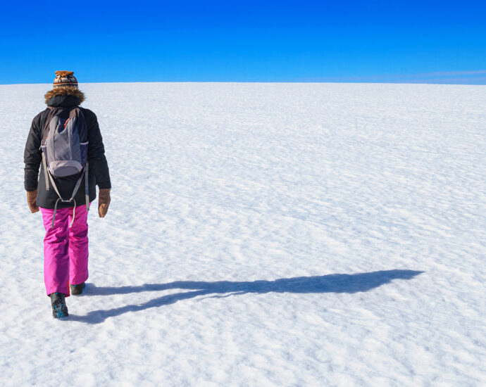 An empty, snow-covered, arctic winter landscape; a snowy desert field with a person walking away toward a blue sky at the horizon.