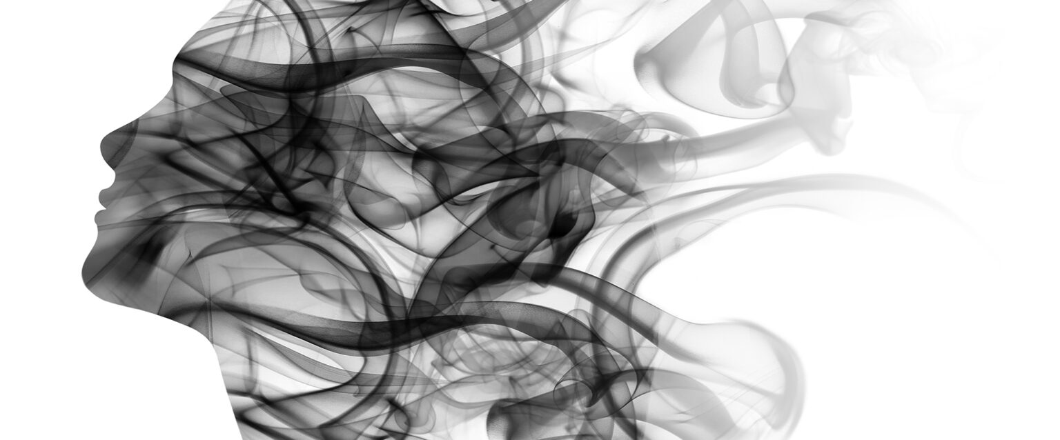 Black smoke in the shape of a side portrait of a woman's face. The black tendrils of smoke create the feeling of long wavy hair blowing in the wind. Double exposure.