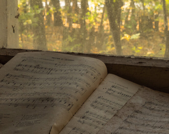 A dirty book of music, with one page torn out, lies open against a window overlooking a wooded area, within an abandoned kindergarten in the Chernobyl Exclusion Zone.