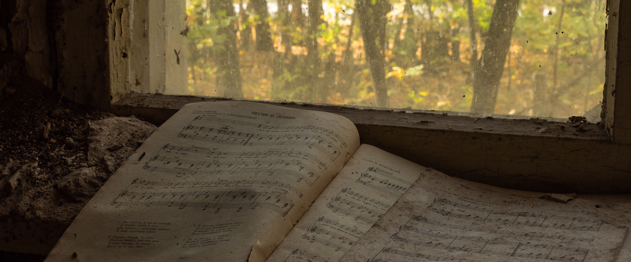 A dirty book of music, with one page torn out, lies open against a window overlooking a wooded area, within an abandoned kindergarten in the Chernobyl Exclusion Zone.