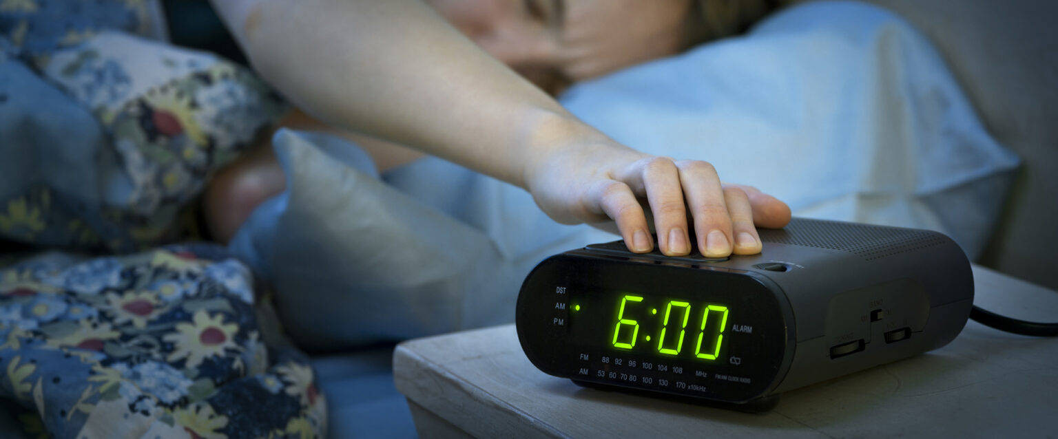 A young woman reaches over, from beneath flowered blankets, to press the snooze button on a black digital alarm clock radio. The green, glowing numbers show that it is 6:00 AM.