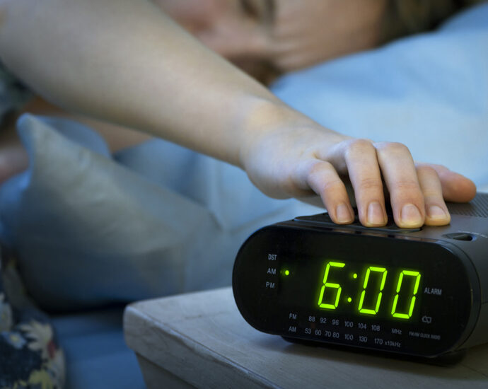 A young woman reaches over, from beneath flowered blankets, to press the snooze button on a black digital alarm clock radio. The green, glowing numbers show that it is 6:00 AM.