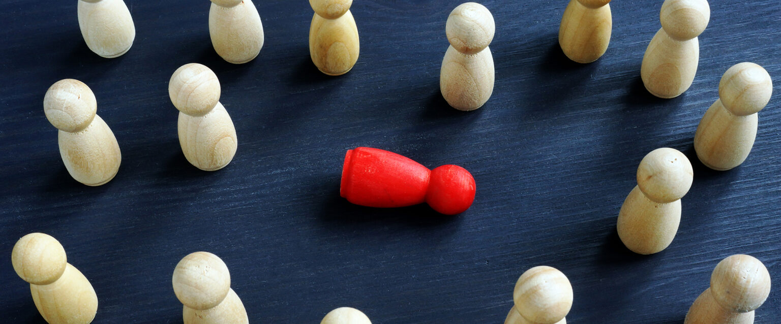 Viewed from above, 15 wooden figurines, made from white pine, stand atop a table. They are surrounding a single red figurine, which is lying on its side amongst them. The image represents bullying, harassment and loneliness.