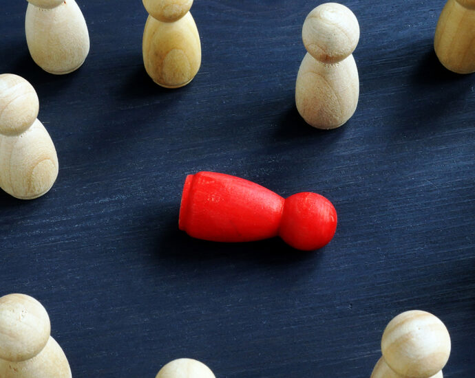 Viewed from above, 15 wooden figurines, made from white pine, stand atop a table. They are surrounding a single red figurine, which is lying on its side amongst them. The image represents bullying, harassment and loneliness.