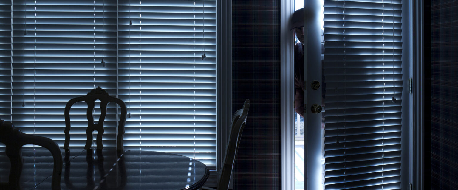 The silhouette of a burglar sneeking into a home, through the kitchen door. View from inside a residence with blinds closed.