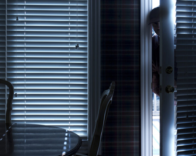 The silhouette of a burglar sneeking into a home, through the kitchen door. View from inside a residence with blinds closed.