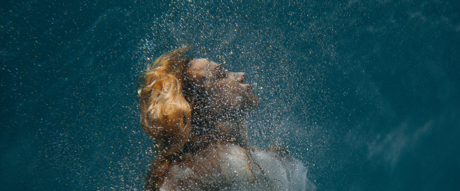 Surrounded by tiny bubbles, a woman in her bridal dress sinks deep beneath the surface of the water.