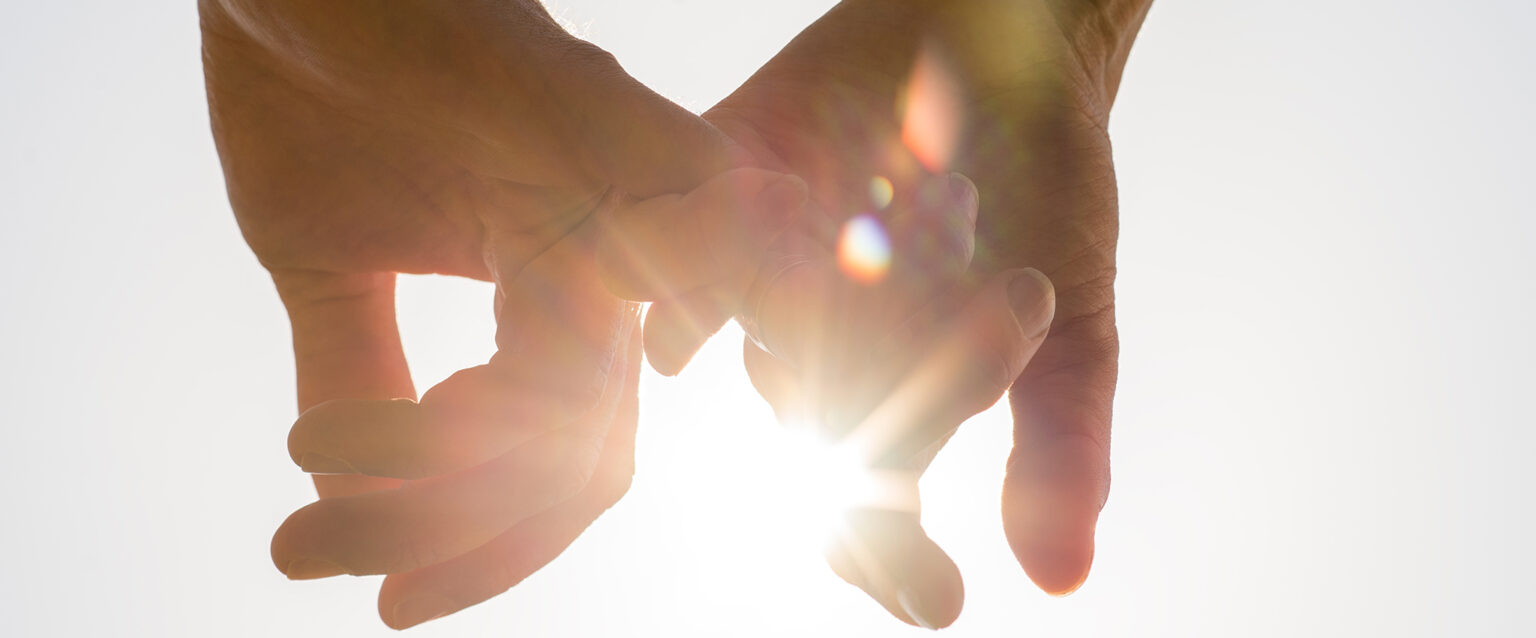 A couple holding hands, facing toward the sun, with a bright sun flare between the silhouetted fingers against a pale blue sky.