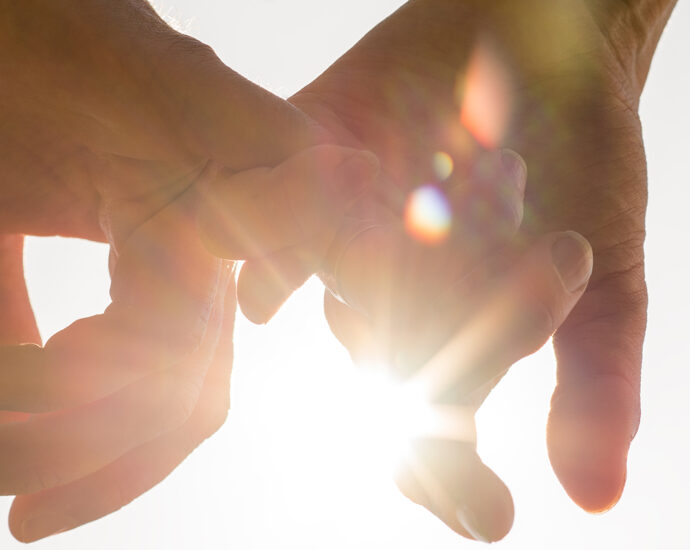 A couple holding hands, facing toward the sun, with a bright sun flare between the silhouetted fingers against a pale blue sky.