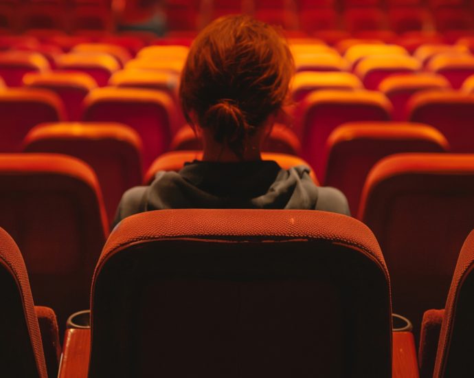 A lonely woman sits alone in the back row of an empty movie theater. The orange/red seats are illuminated, from above, by a yellow light.