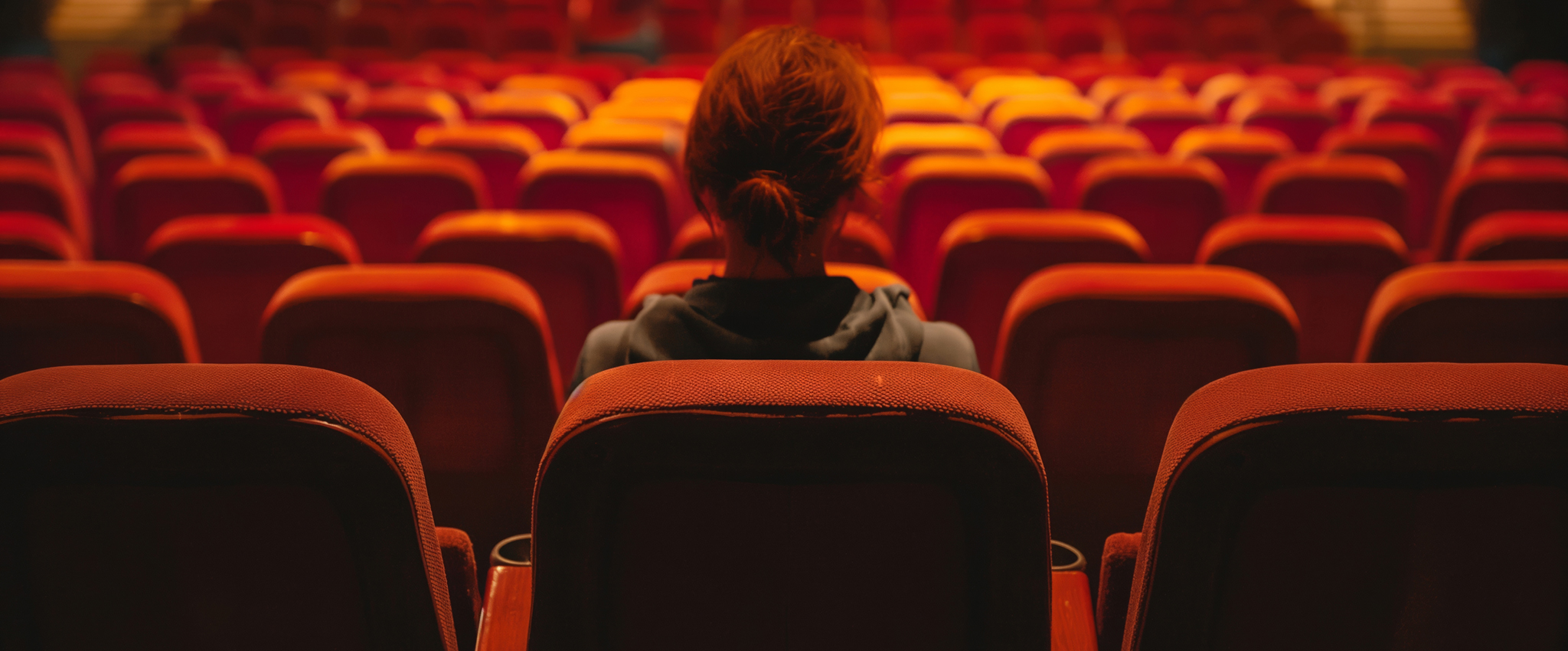 A lonely woman sits alone in the back row of an empty movie theater. The orange/red seats are illuminated, from above, by a yellow light.