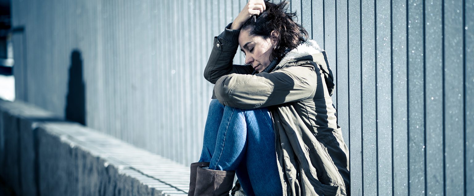 A sad, young woman sits on a cement ledge, contemplating. Abusive relationship and harassment concept