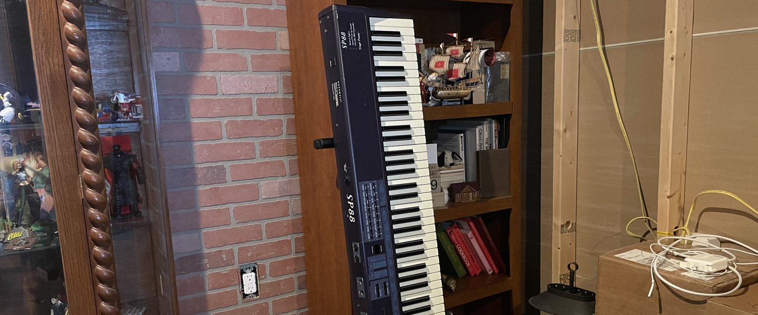 IMG_2258-2048x850 | E.L. Redwine Two stacked electronic keyboards on a wooden shelf against a red brick wall, with ship models and books on the shelves nearby.