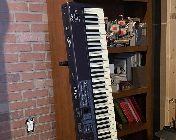 Two stacked electronic keyboards on a wooden shelf against a red brick wall, with ship models and books on the shelves nearby.